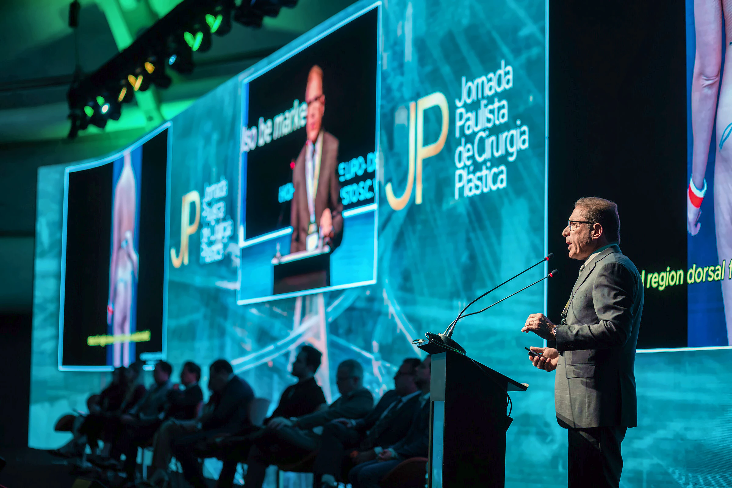 Dr. Augusto Valente palestrando ao púlpito diante de plateia de cirurgiões, com telões ao fundo, na Jornada Paulista de Cirurgia Plástica